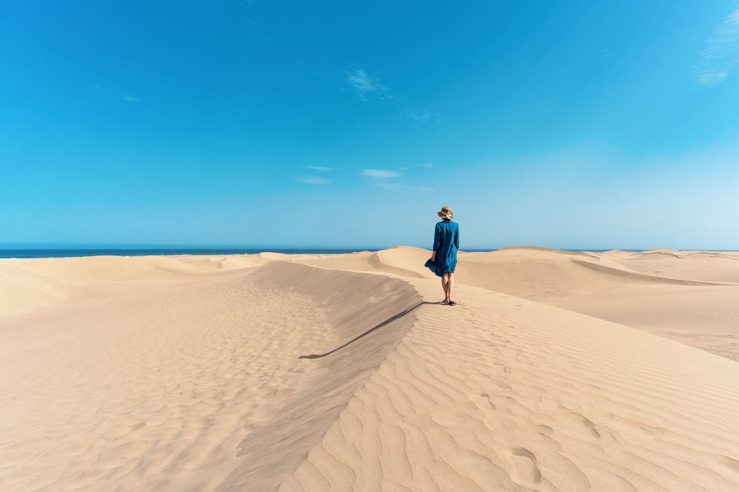 Una mujer con sombrero de paja y vestido azul camina por la cresta de una duna junto al mar.